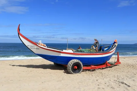 En traditionell blåvit fiskebåt med högt uppsvängd stäv står på en vagn i strandsanden medan två fiskare gör i ordning tåg och nät ombord