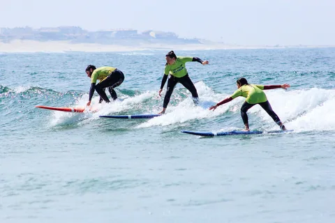 Tre surfelever i likadana gulgröna tröjor som reser sig på mjuka nybörjarbrädor i en liten våg nära stranden