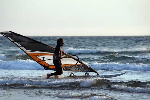 En vindsurfare med uppriggat segel glider genom brytande vitt vatten nära stranden