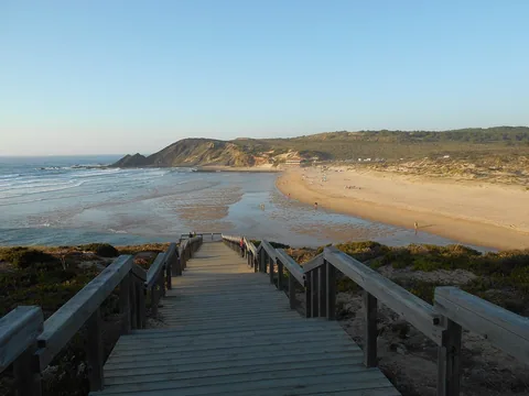 Träspång som leder ner mot en bred och glest besökt sandstrand med en klippudde vid strandkanten och Atlanten bortom