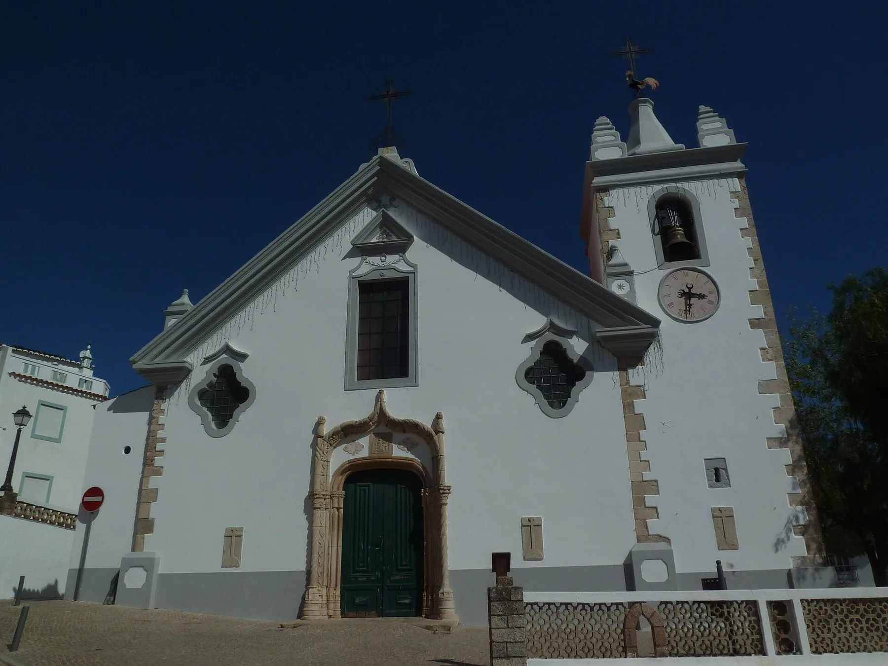 Vitkalkad bykyrka med snidad stenportal och klocktorn med kyrkklocka och urtavla mot djupblå himmel