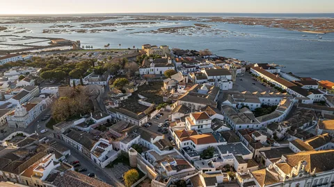 Flygvy över en gammal stadskärna med vitkalkade hus och tegeltak innanför stadsmuren, med en grund lagun och sandbankar bakom