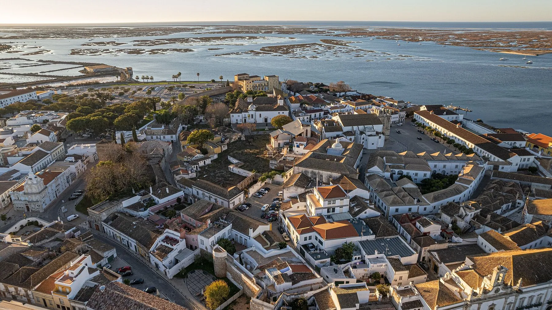 Flygvy över en gammal stadskärna med vitkalkade hus och tegeltak innanför stadsmuren, med en grund lagun och sandbankar bakom