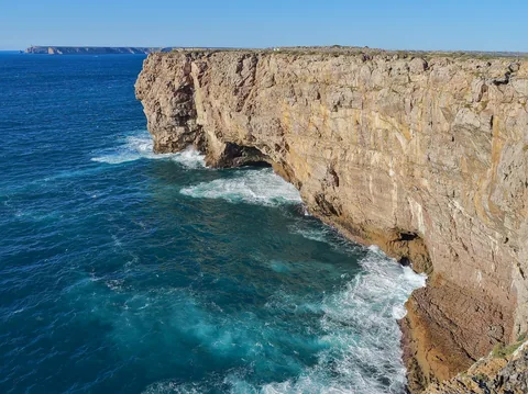 Lodrät skiktad klippvägg som faller rakt ner i turkost hav vid Sagres, med bränning mot foten
