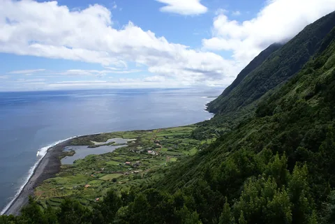 En brant grönklädd bergssida faller rakt ned mot en låg kustplatå med en lagun, några vita hus och en svart strandkant
