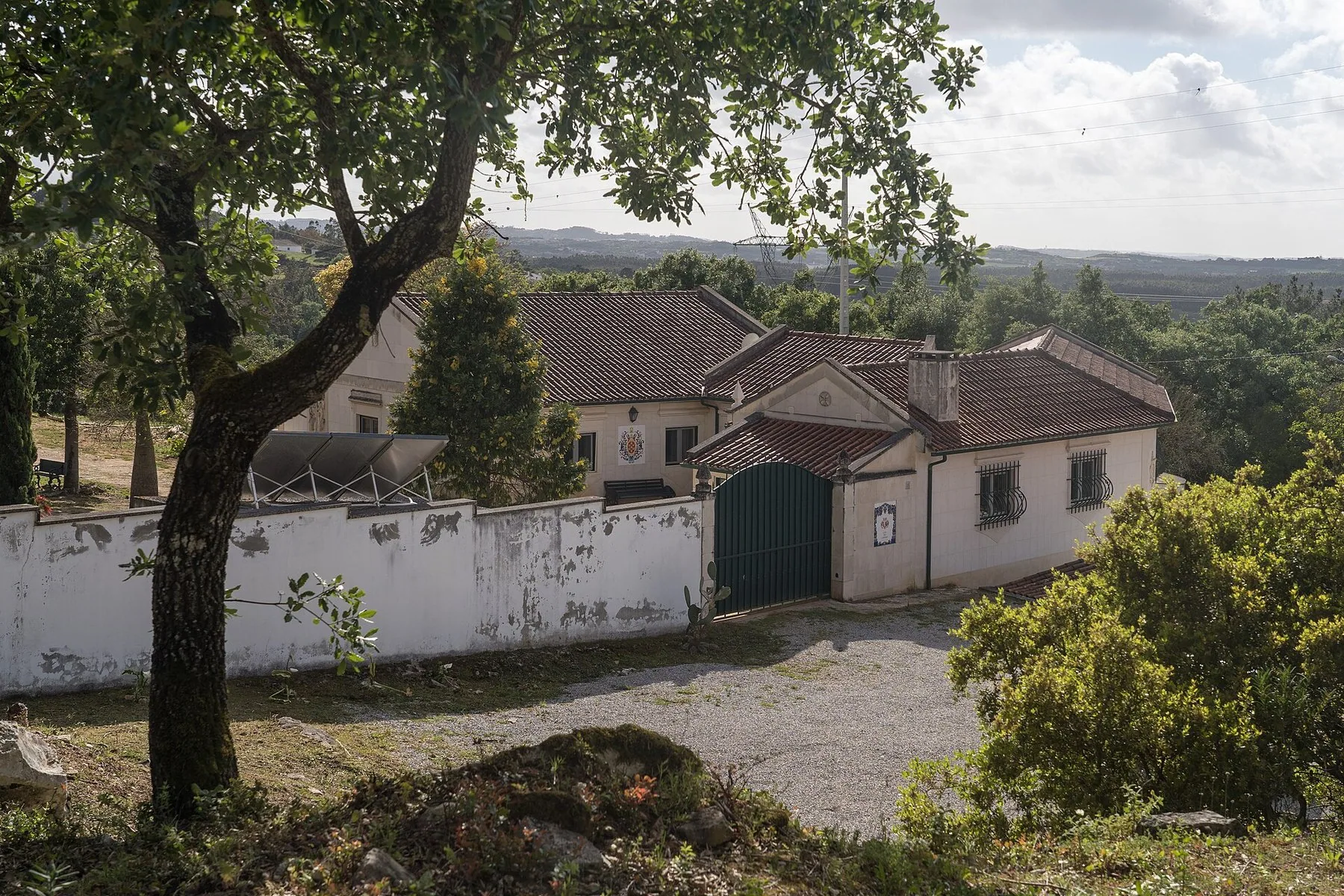 En vitkalkad gård med tegeltak och grön port bakom en låg mur, omgiven av fruktträd med kullar och skog i bakgrunden