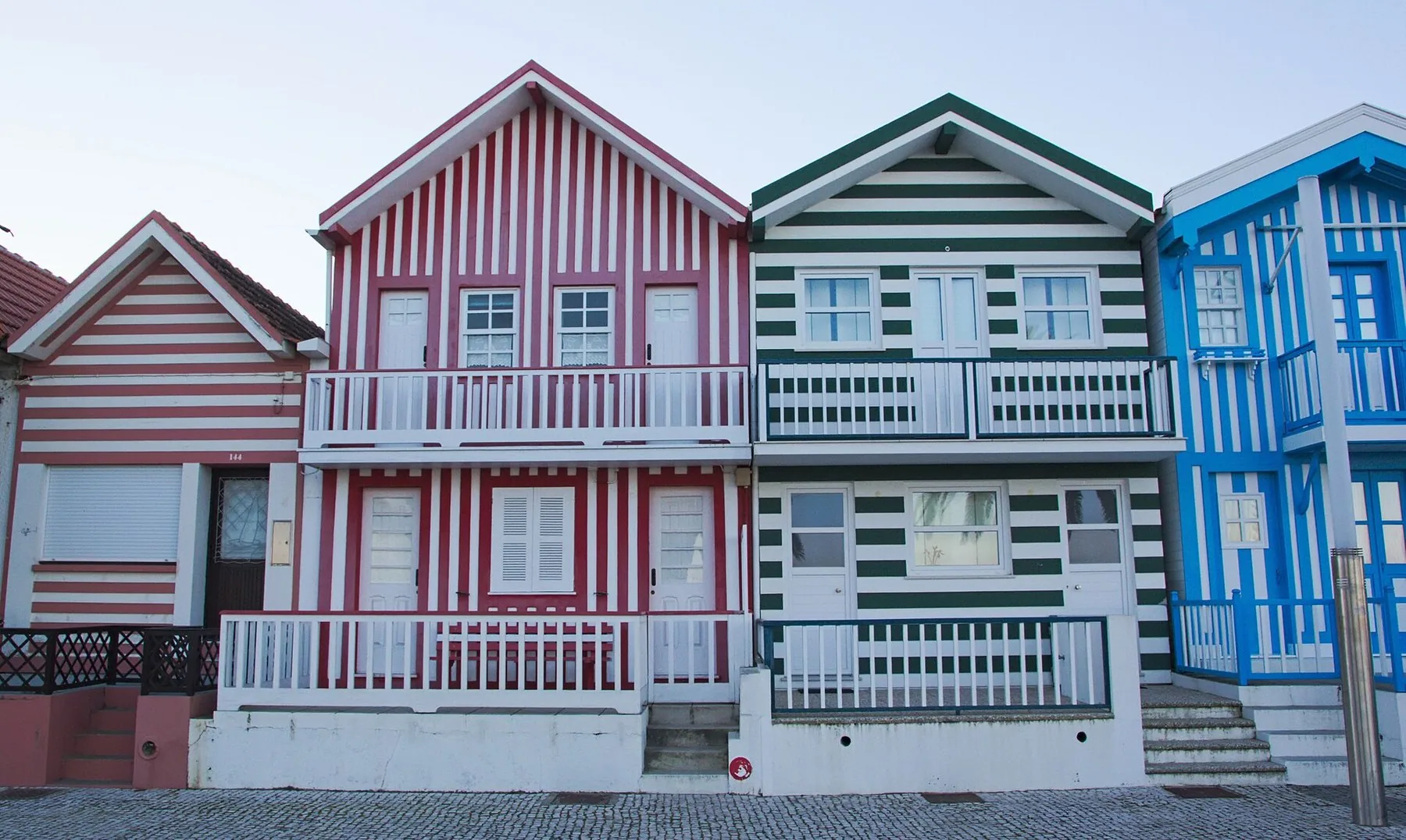 Rad av trähus i Costa Nova med lodräta ränder i rosa, rött, grönt och blått och vita verandaräcken mot gatan