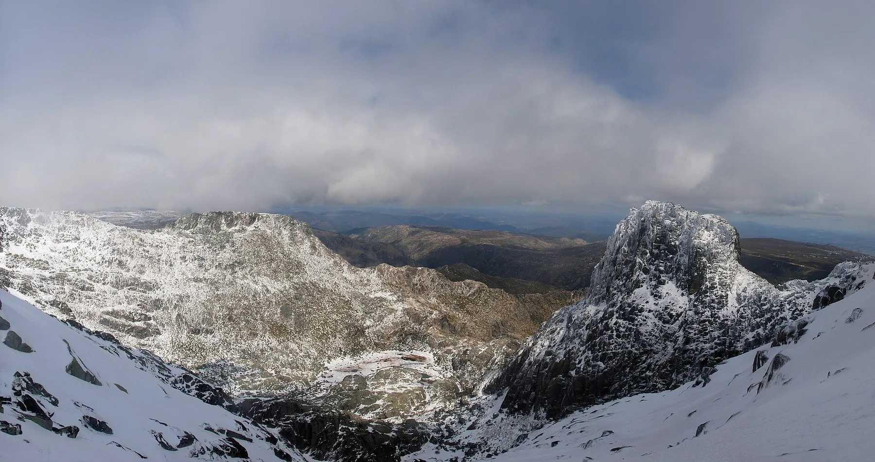 Snöklädda bergssidor och en brant klippformation i Serra da Estrela under tunga moln