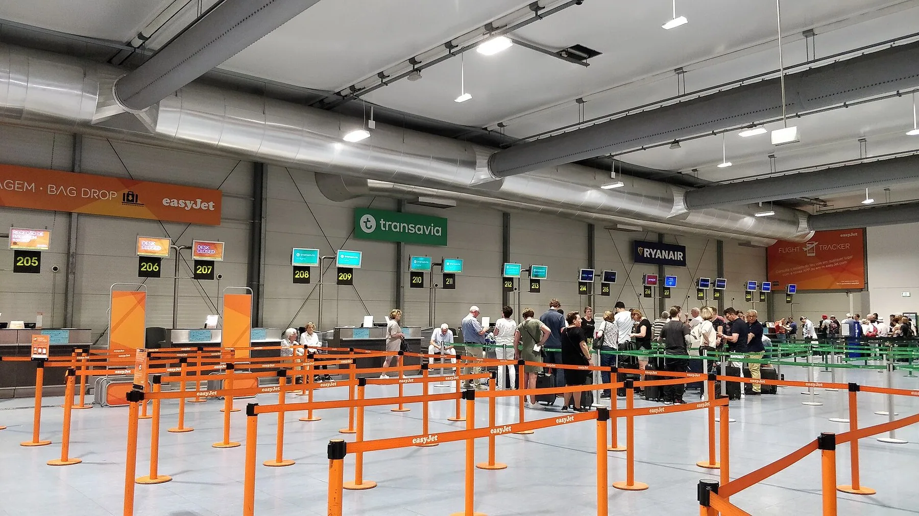 Incheckningshall med bag drop-diskar för easyJet, Transavia och Ryanair, avgränsade av orange köbandsstolpar, och en kö av resenärer med väskor längst bort