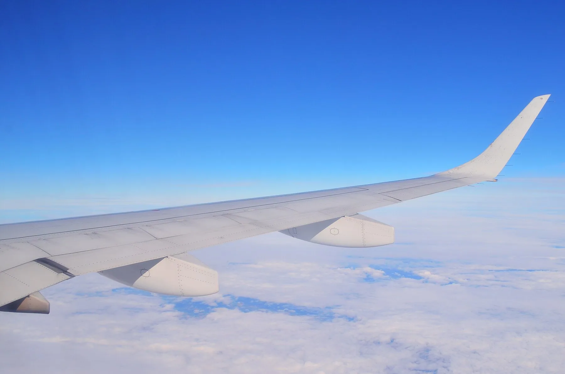 Ett flygplans vinge med uppåtböjd vingspets över ett vitt molntäcke under klarblå himmel