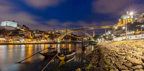Dom Luís I-bron och den upplysta flodstranden i Porto i blå timme, med rabelobåtar på Douro
