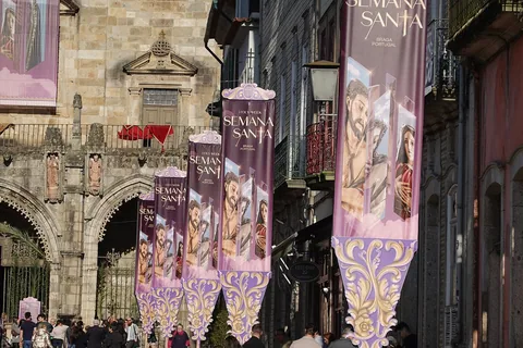 Höga lila banderoller med texten Semana Santa hänger utmed en smal gata framför en stenkyrkas portal, med folk samlade nedanför