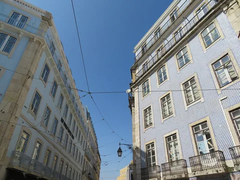 Två pombalinska husfasader sedda underifrån mot blå himmel, den ena klädd i blåvita kakelplattor, båda med smidesbalkonger och en gatlykta på en spårvagnstråd mellan sig