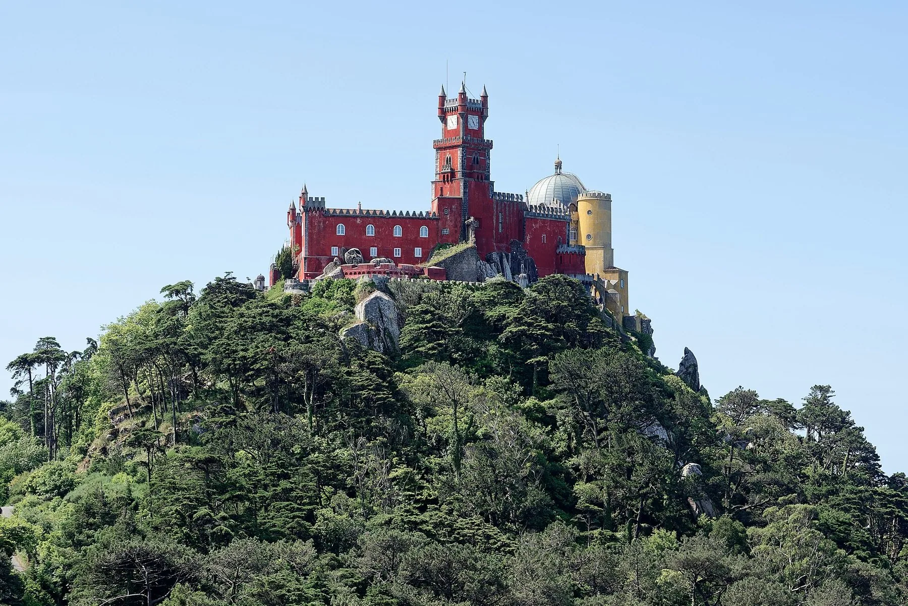 Palácio da Pena i rött och gult kröner en tätt skogklädd bergstopp under klarblå himmel