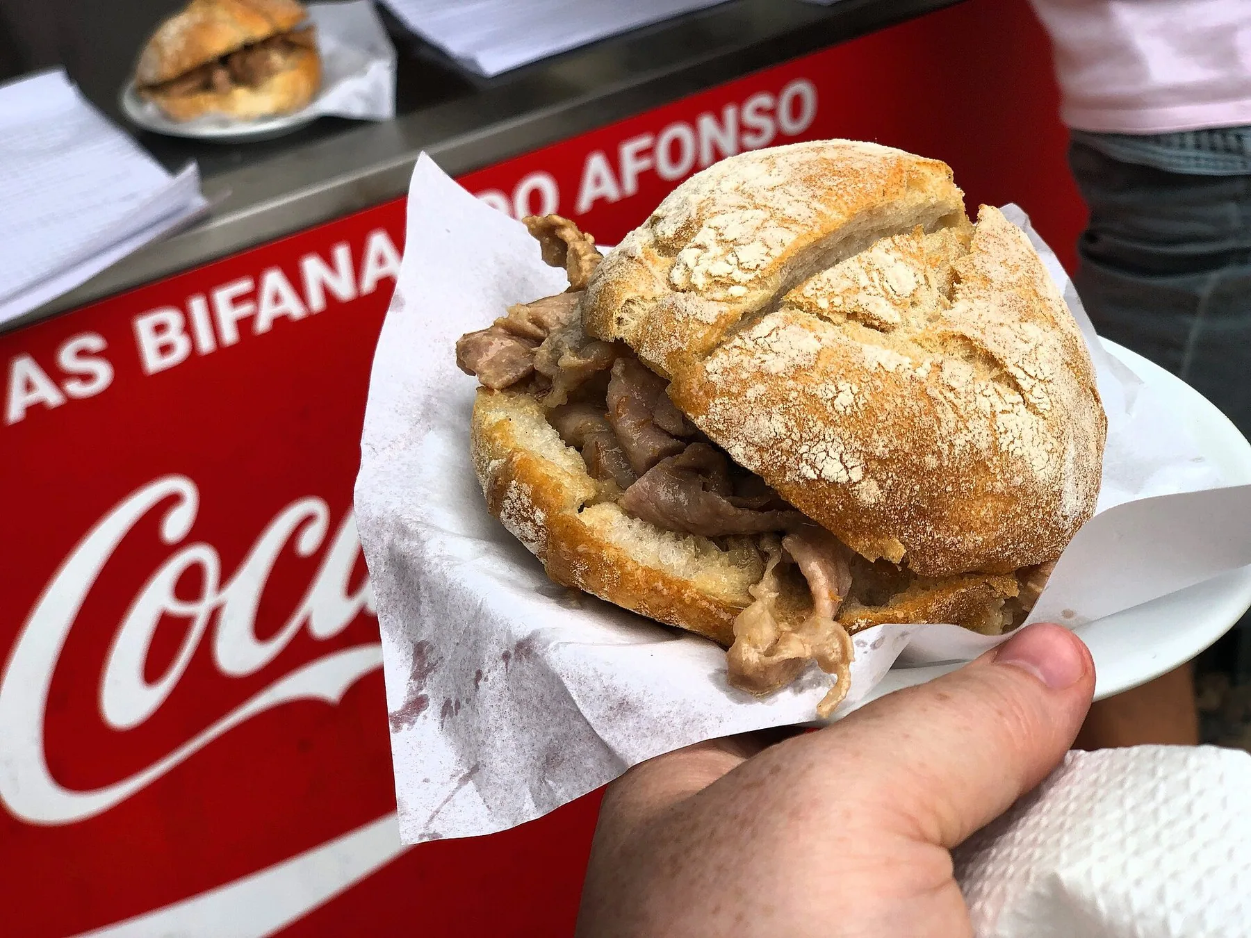 En bifana i mjöligt portugisiskt bröd med tunna skivor stekt fläsk som sticker ut i kanterna, hållen i handen på ett serveringspapper