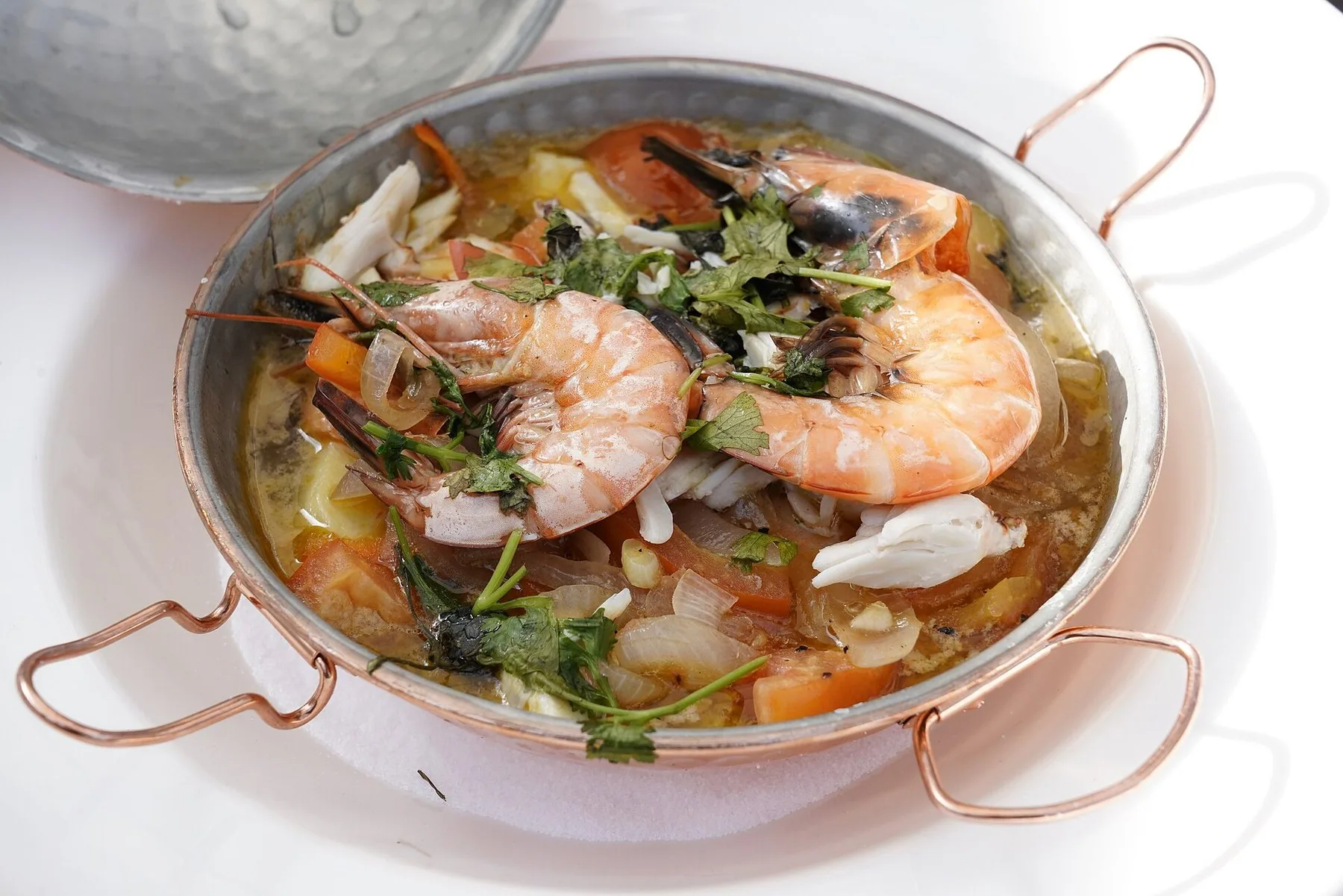 Öppnad kopparcataplana med räkor, fisk och tomat i buljong, med kärlets böjda handtag runt kanten