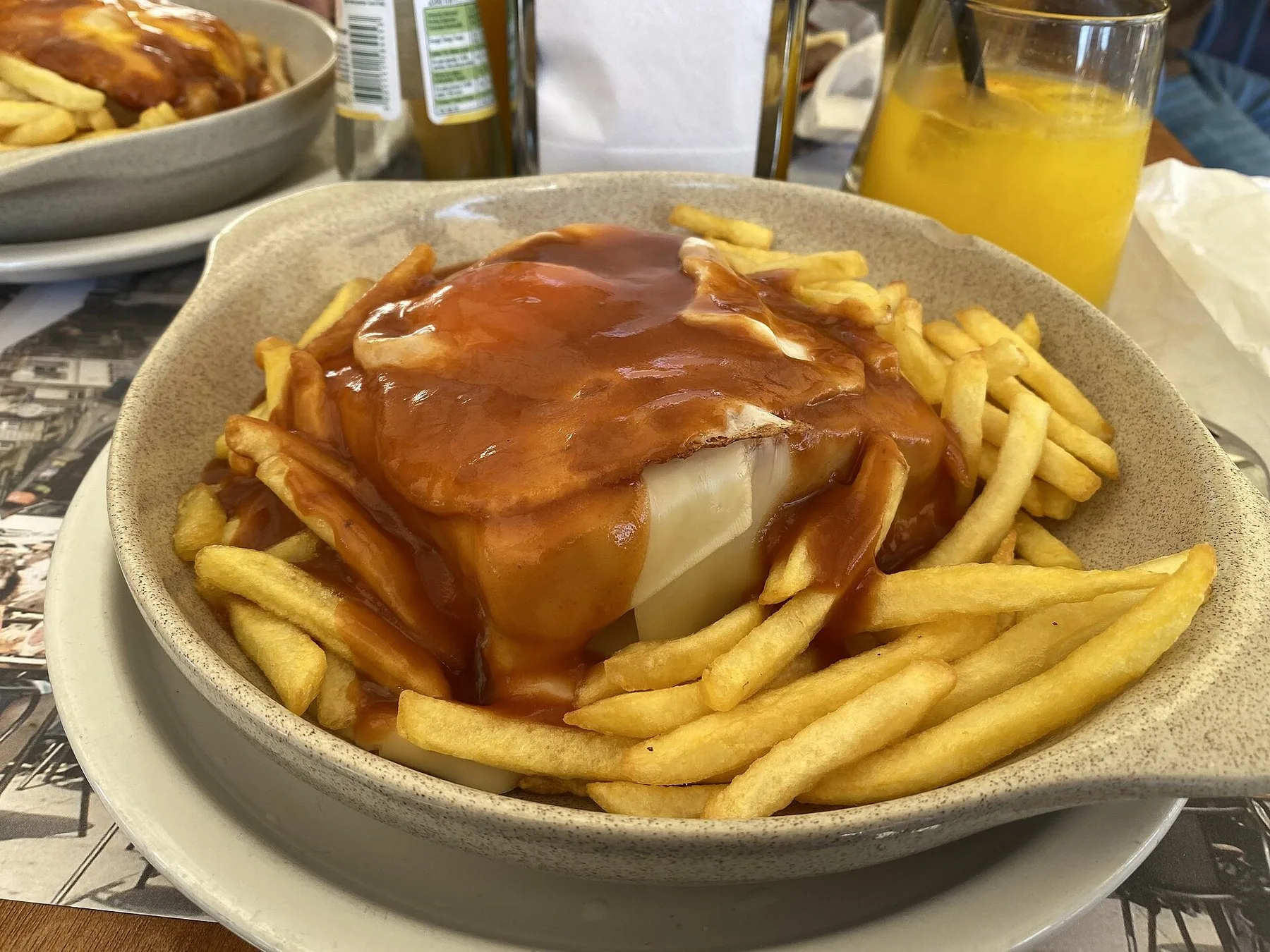 Francesinha med smält ost och tjock sås i en djup skål, omgiven av pommes frites