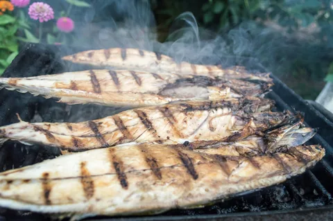 Fyra hela fiskar med grillränder ligger på ett galler medan röken stiger