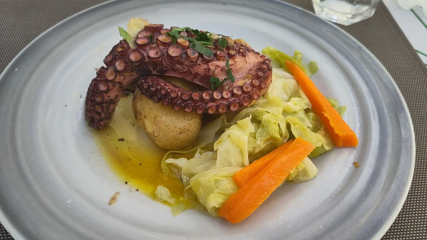 En hel bläckfiskarm över en kokt potatis på tallriken, med kål och morot bredvid och en glänsande sås av olivolja runt om