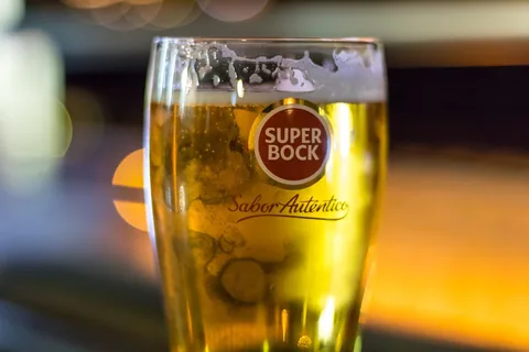 Ett kylslaget glas ljus fatöl med skumkrona i ett Super Bock-glas, mot suddiga varma kaféljus i bakgrunden