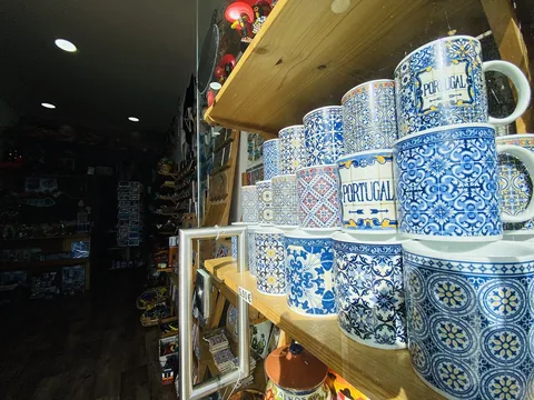 Staplade blåvitmönstrade muggar märkta Portugal på en butikshylla, med prislapp framför och souvenirställ längre in i den mörka lokalen