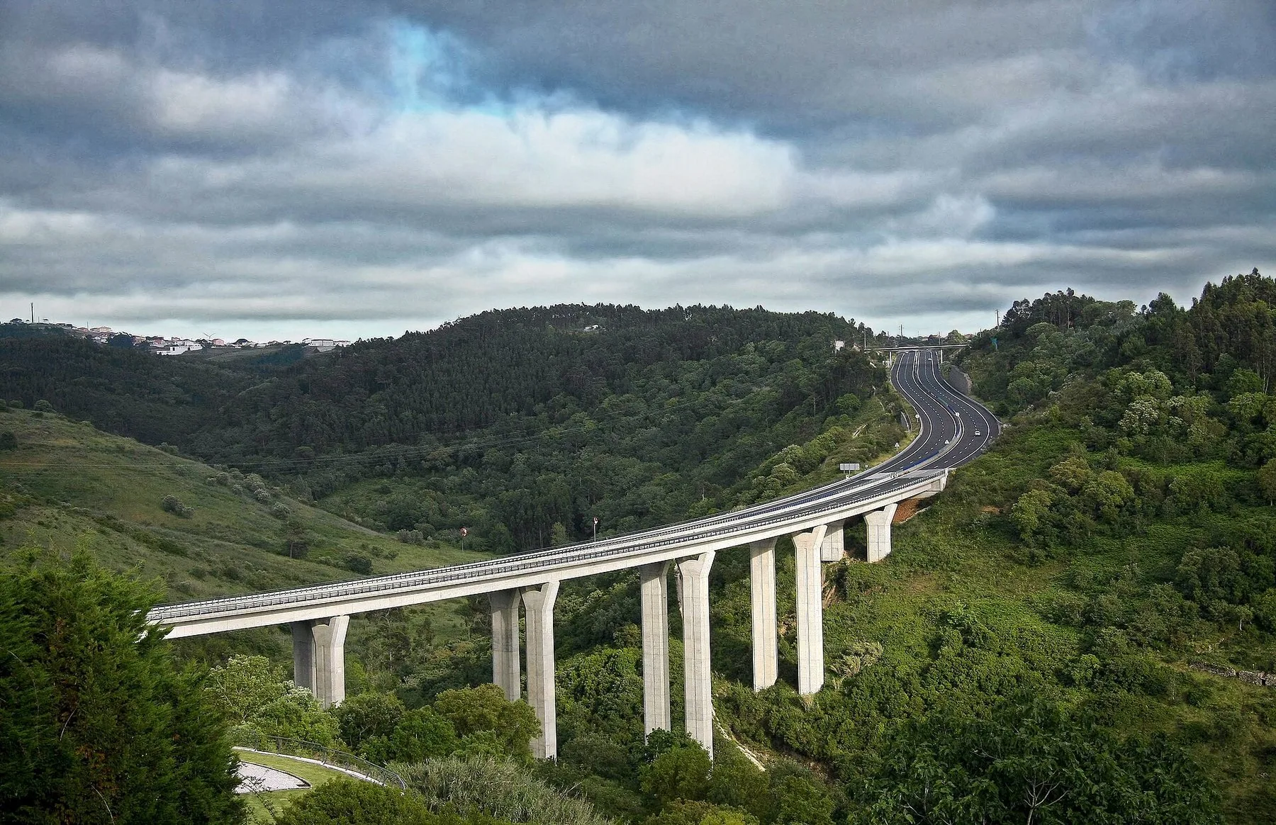 En motorvägsviadukt på höga pelare svänger fram genom en grön dalgång med skogklädda kullar under en molnig himmel