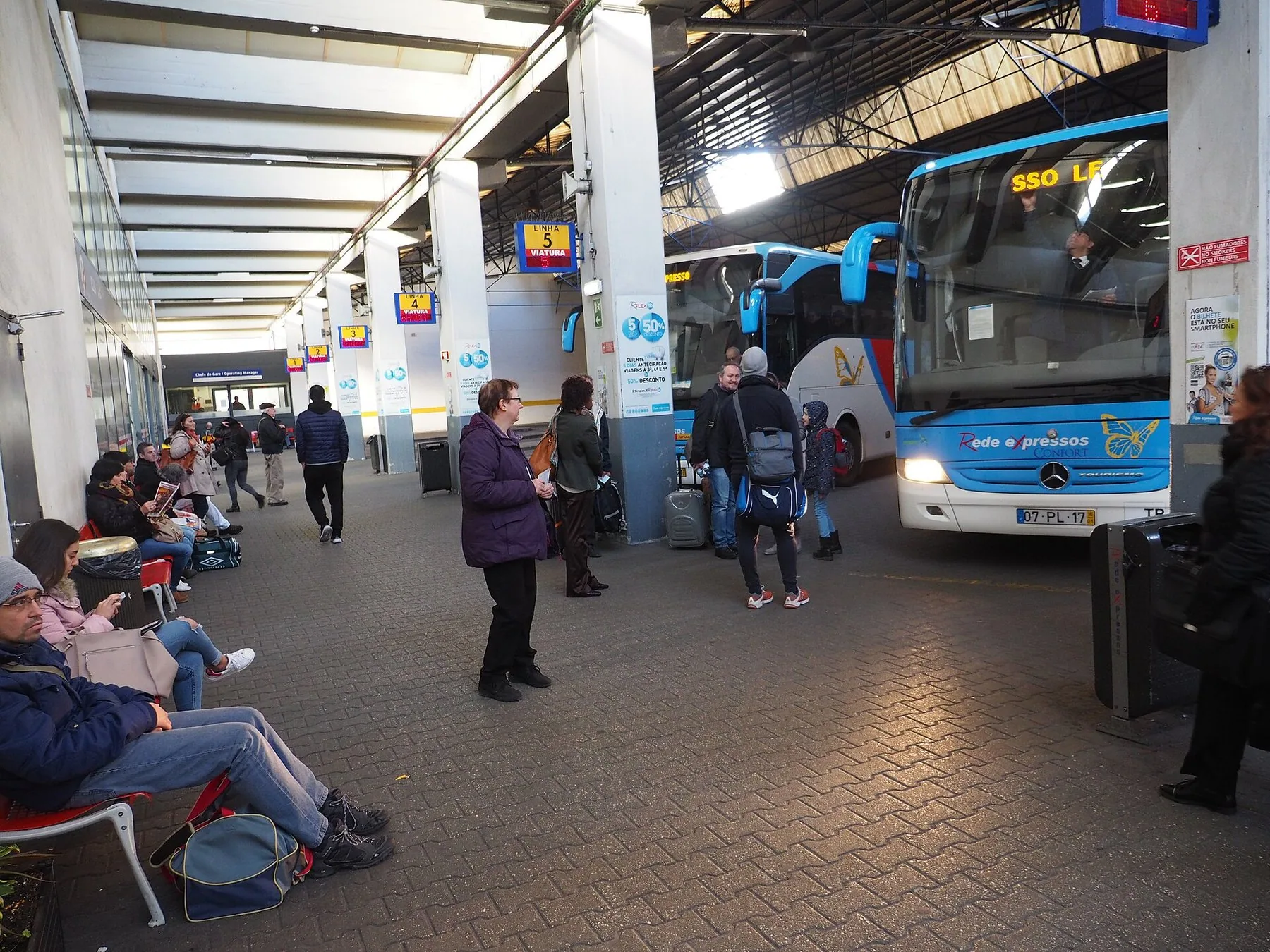 Resenärer med väskor väntar under ett bussterminaltak medan en långfärdsbuss står med tända lyktor vid perrong fem