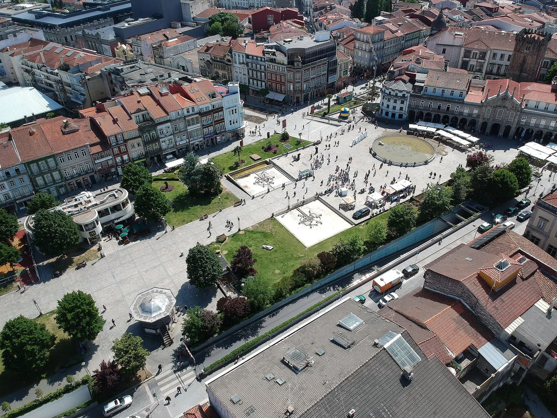 Stort torg i en stadskärna sett uppifrån, med fontäner, trädrader och tegeltak tätt runt omkring