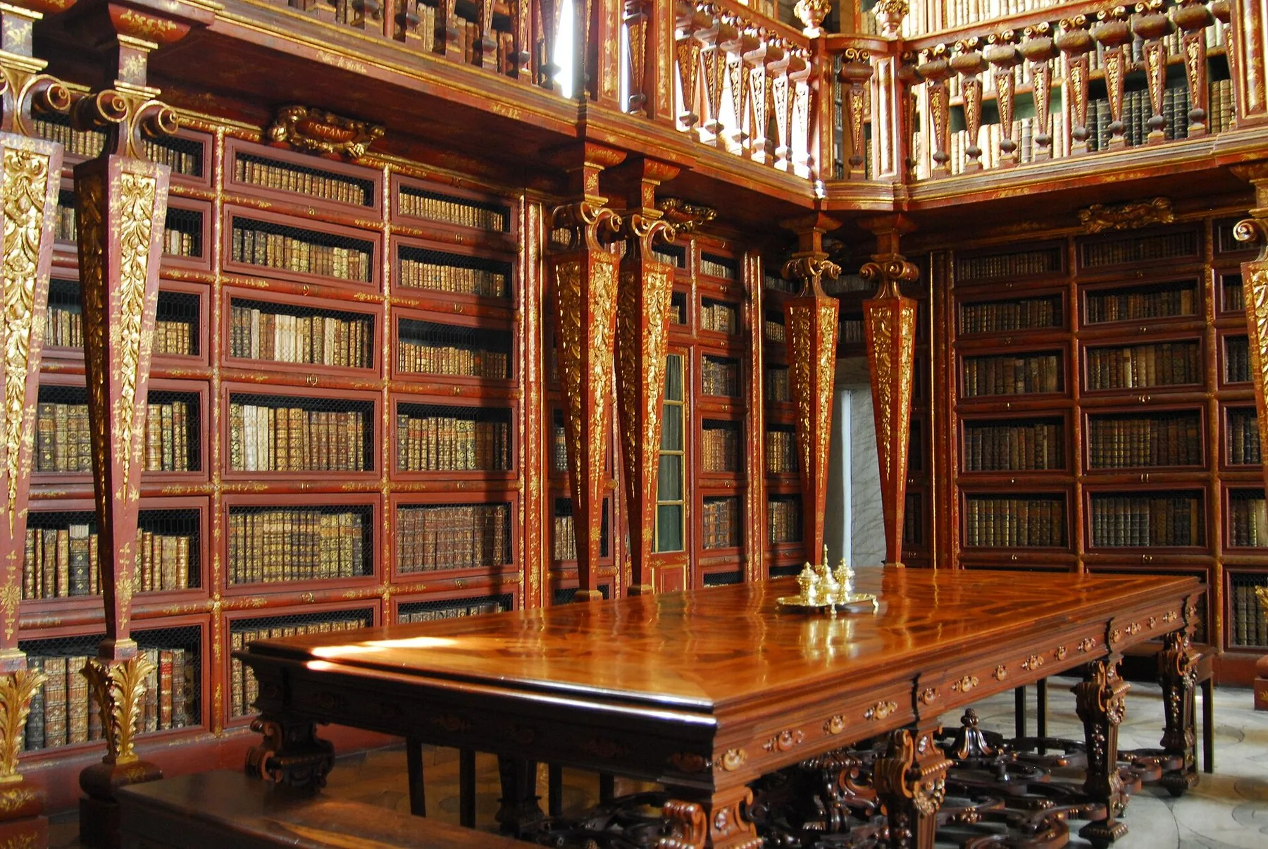 Barocksalen i Biblioteca Joanina med bokhyllor i mörkt trä och förgyllda ornament i två våningar, och ett långt blankt träbord längs mittgången