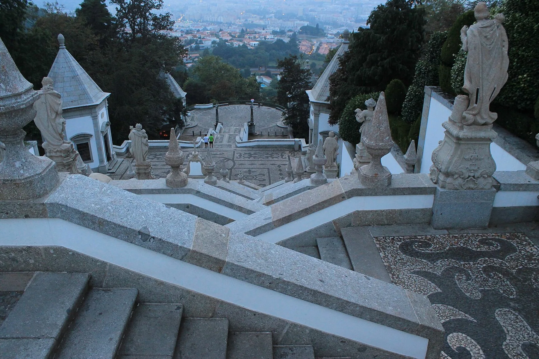 Barocktrappan vid Bom Jesus do Monte sedd uppifrån: vita trapplopp i sicksack med granitstatyer och stenspiror, och staden Braga i diset nedanför