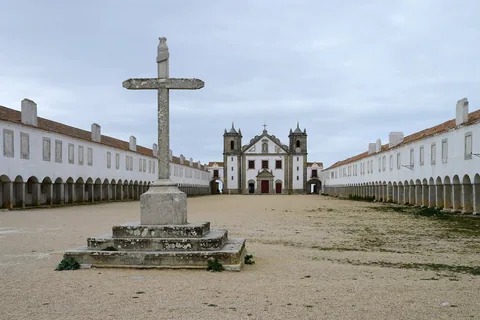 Ett stenkors på trappsockel mitt på en tom grusgård mellan två långa arkadflyglar som möts i en vitkalkad kyrka med två torn