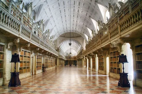 Biblioteket i Mafra: en lång tunnvälvd sal i vitt och guld med bokhyllor i två våningar längs väggarna och rutmönstrat marmorgolv