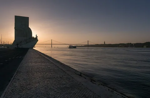 Padrão dos Descobrimentos i siluett från sidan vid soluppgången, format som ett skeppsstäv med raden av figurer, med Tejo och hängbron bakom