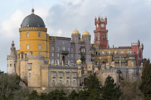 Penaslottet på bergstoppen i Sintra med gul kupol, röda torn och krenelerade murar mot molnig himmel