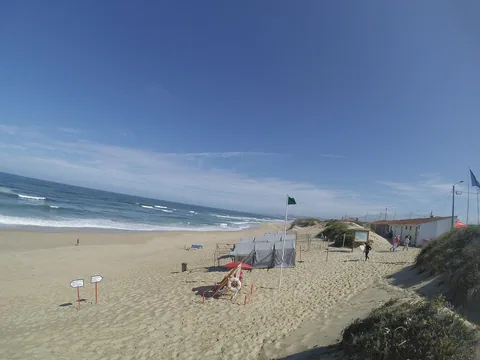 Grön flagga på en flaggstång vid strandkanten intill en badvaktspost med livboj och räddningsbräda, medan vågorna bryter i flera rader utanför.
