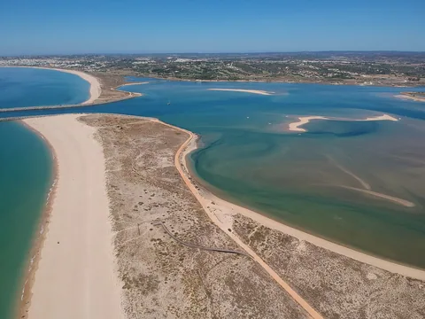 Flygbild över Ria de Alvor: en smal sandrevel skiljer det öppna havet från den grunda lagunen innanför, där sandbankar ligger blottade i det turkosa vattnet.