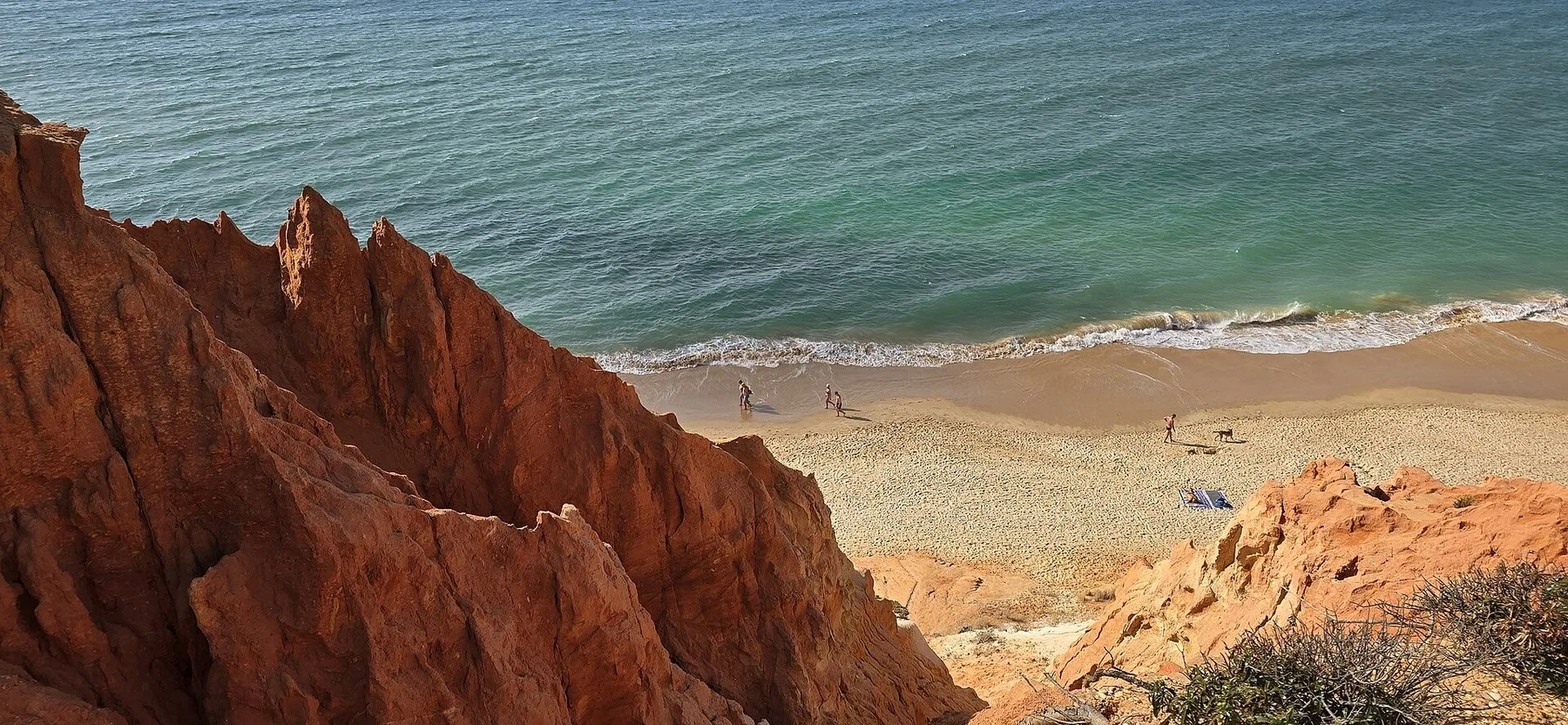 De rödorange, djupt eroderade klipporna vid Praia da Falésia faller ner mot en långsträckt sandstrand där vågorna bryter längs vattenlinjen.