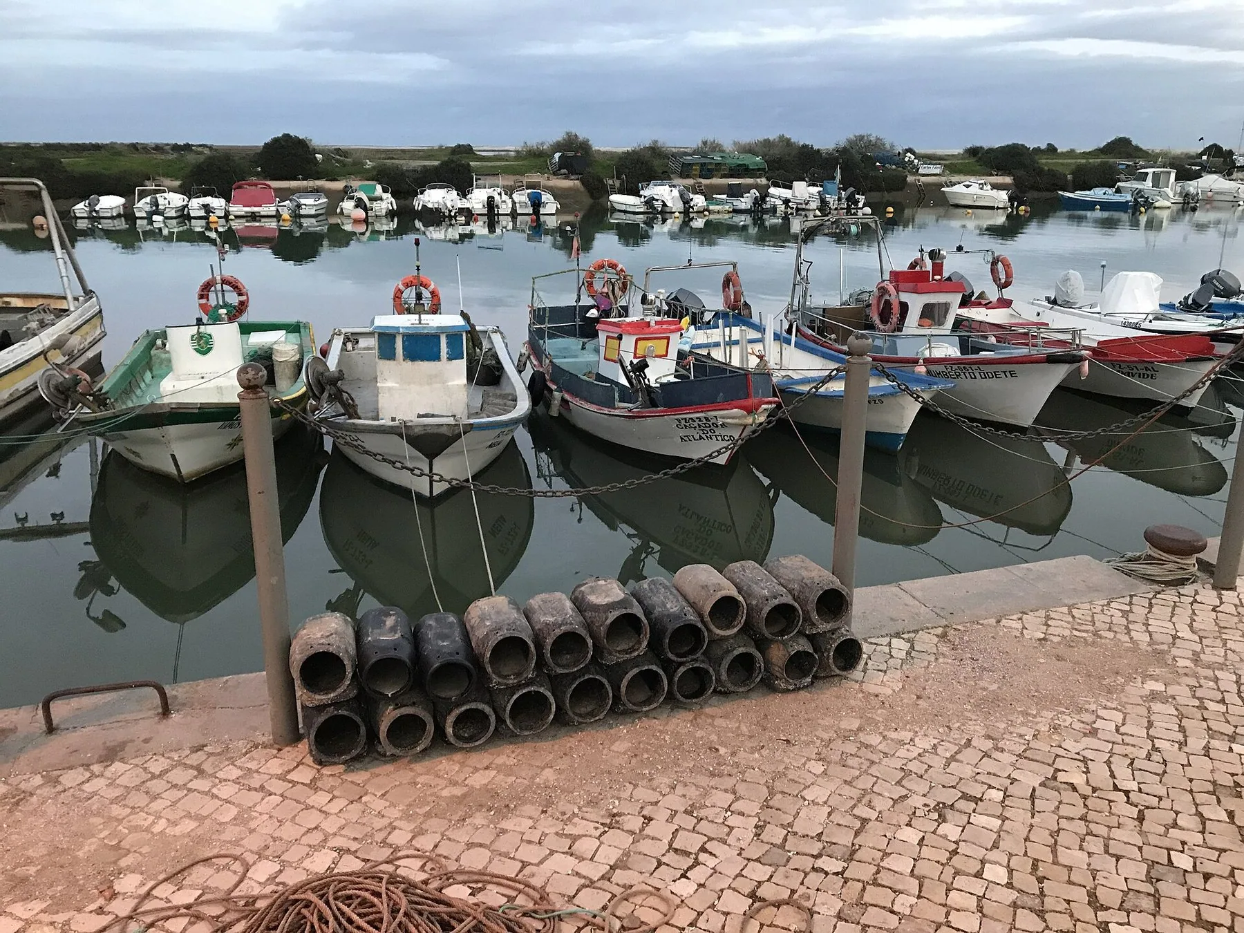 Små färgade fiskebåtar förtöjda i rad längs kajen i Fuseta, speglade i stilla vatten