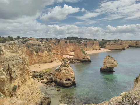 Praia da Marinha sedd från klippkanten: gulbruna kalkstensväggar ramar in en nästan tom sandvik, och två fristående klippelare står i det grönskimrande vattnet utanför.