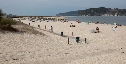 En mycket bred och nästan vit sandstrand med rader av solsängar och glest med badgäster, och tvärs över det smala sundet reser sig en grön bergskam