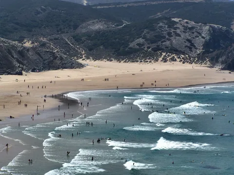 Rad efter rad av bränningar rullar in mot en bred sandstrand där dussintals surfare ligger och väntar ute i vattnet, och bakom stranden reser sig kala, buskbevuxna kullar