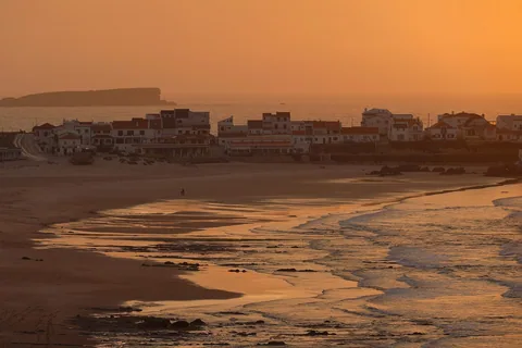 Praia Baleal Norte i kvällsljus: den breda stranden i förgrunden, husraden ute på det smala näset och Baleals klipphalvö som siluett längst bort.