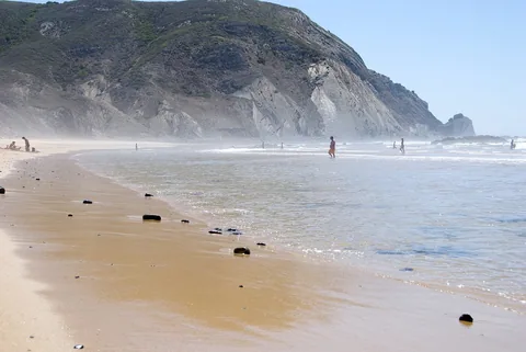 Praia do Castelejo: grov sand med mörka stenar längs vattenbrynet, bränning som rullar in och den höga mörka klippväggen som reser sig i bakgrunden.