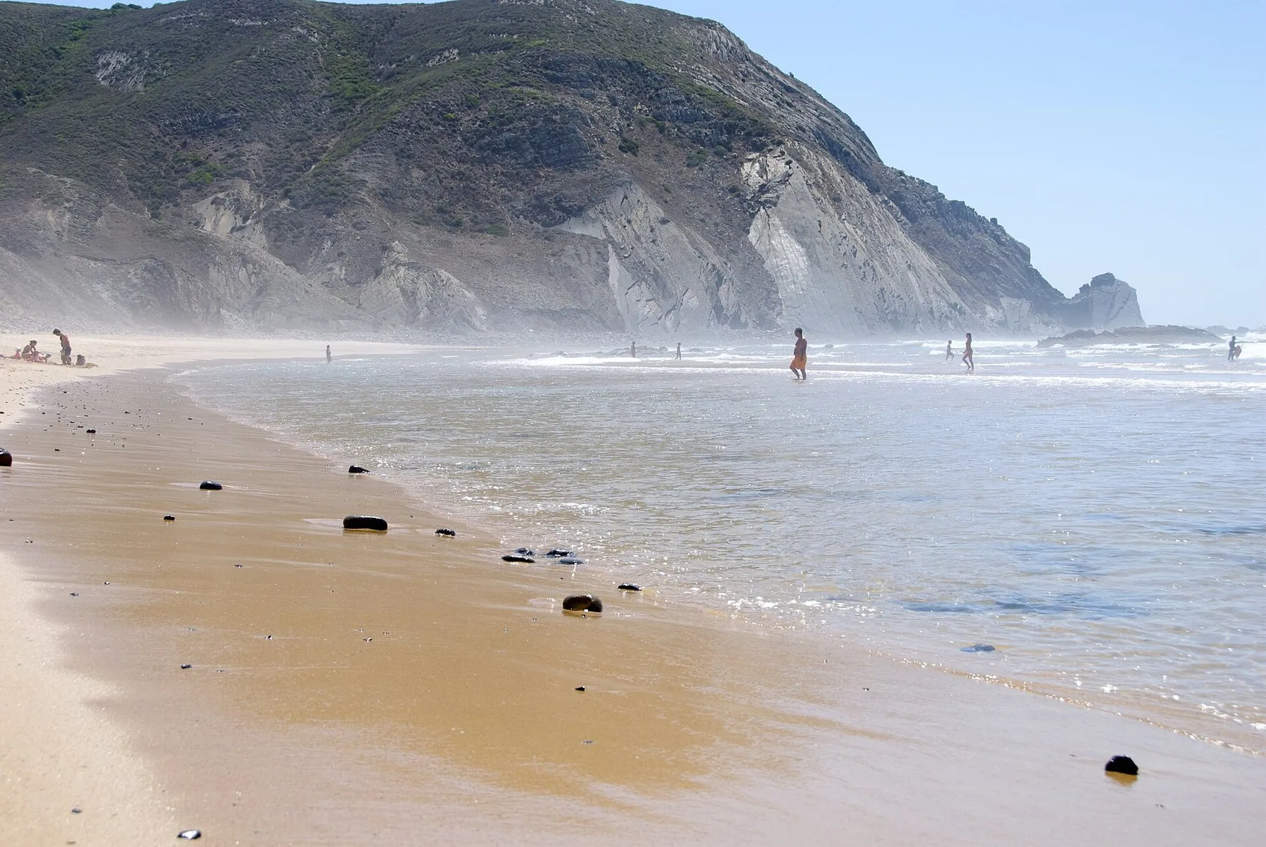 Praia do Castelejo: grov sand med mörka stenar längs vattenbrynet, bränning som rullar in och den höga mörka klippväggen som reser sig i bakgrunden.