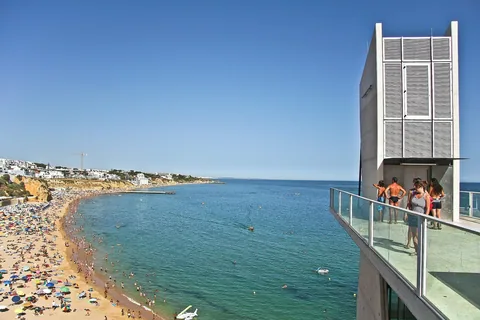 Elevador do Peneco i Albufeira sedd från utsiktsplattformen: hisstornet i betong till höger och nedanför klippan stadsstranden, tätt packad med parasoller och badgäster.