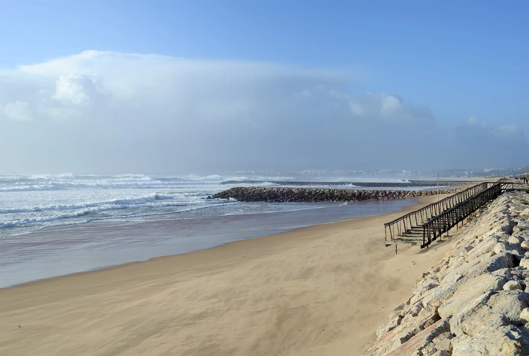 Costa da Caparica: en bred sandstrand med stenvågbrytare som sticker ut i det brytande vattnet, och en trätrappa ner över stenskoningen till höger.
