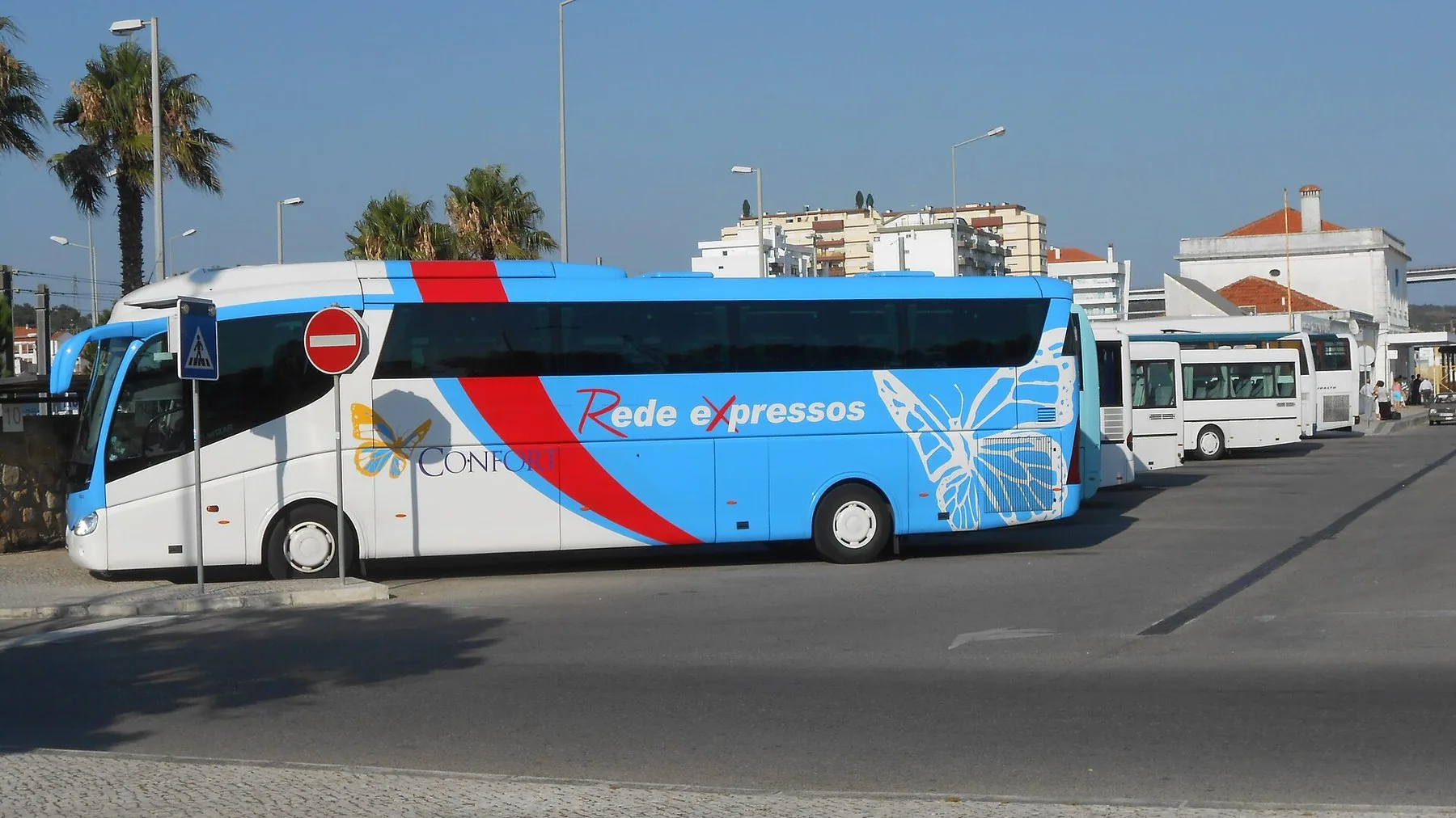 En vit och blå långfärdsbuss märkt Rede Expressos står vid en busshållplats med palmer och låga hus bakom