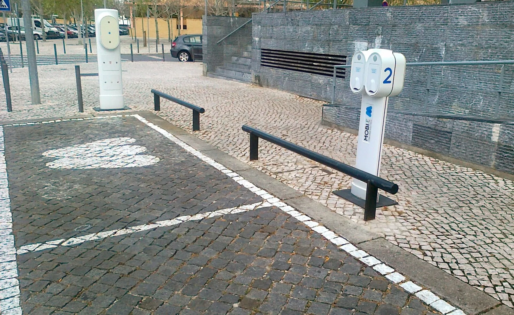 Två vita laddstolpar för elbil på portugisisk kullersten, med vitmålade laddplatser och en blå parkeringsskylt bredvid