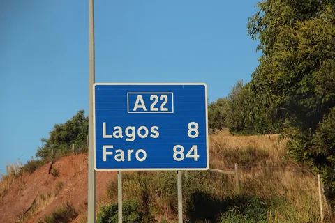 Blå vägskylt vid motorvägen A22 som anger Lagos 8 och Faro 84 kilometer, uppsatt vid en rödbrun slänt med buskar