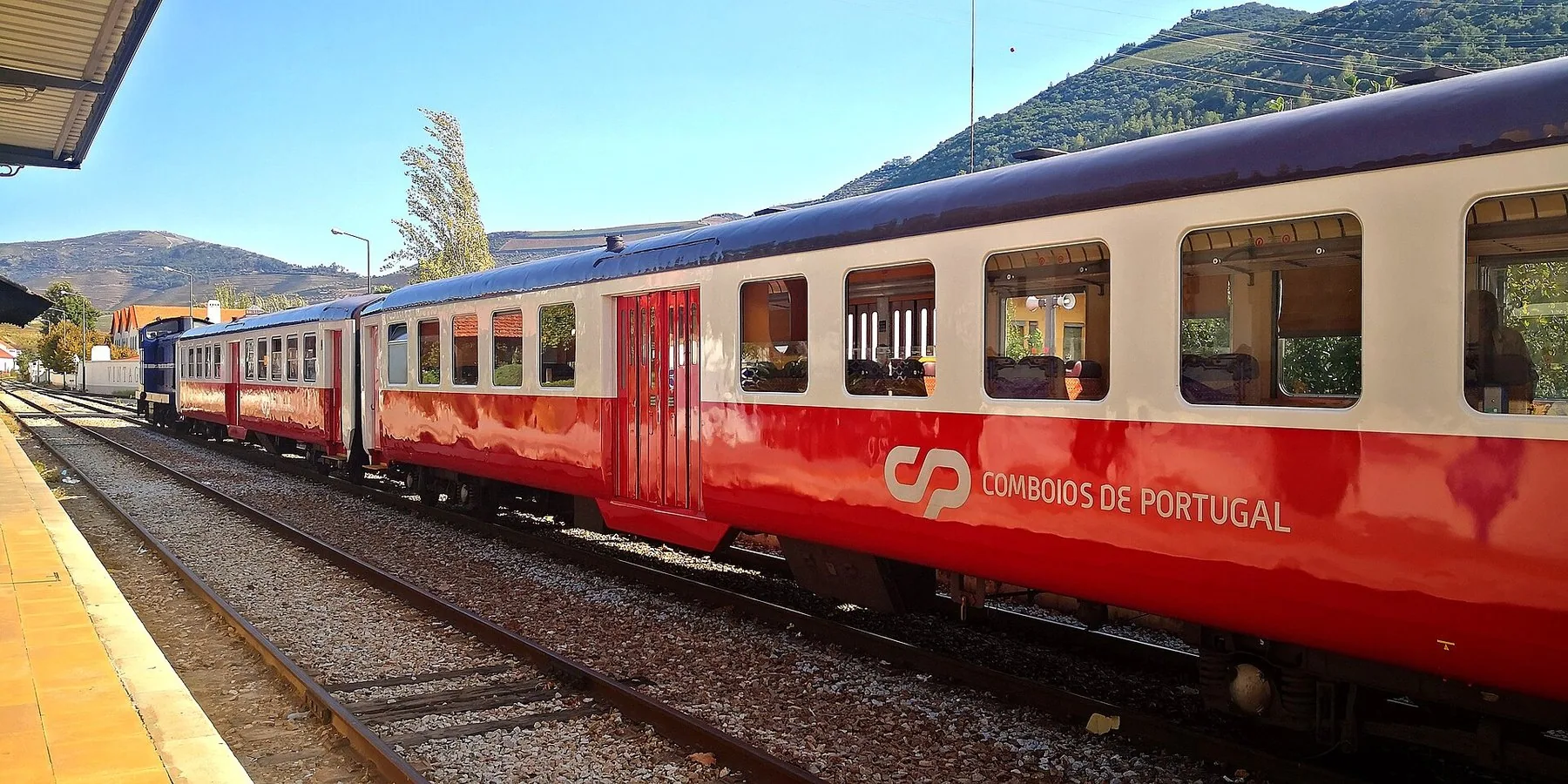 Ett rött och gräddvitt tåg märkt CP Comboios de Portugal står vid en perrong med gröna berg bakom